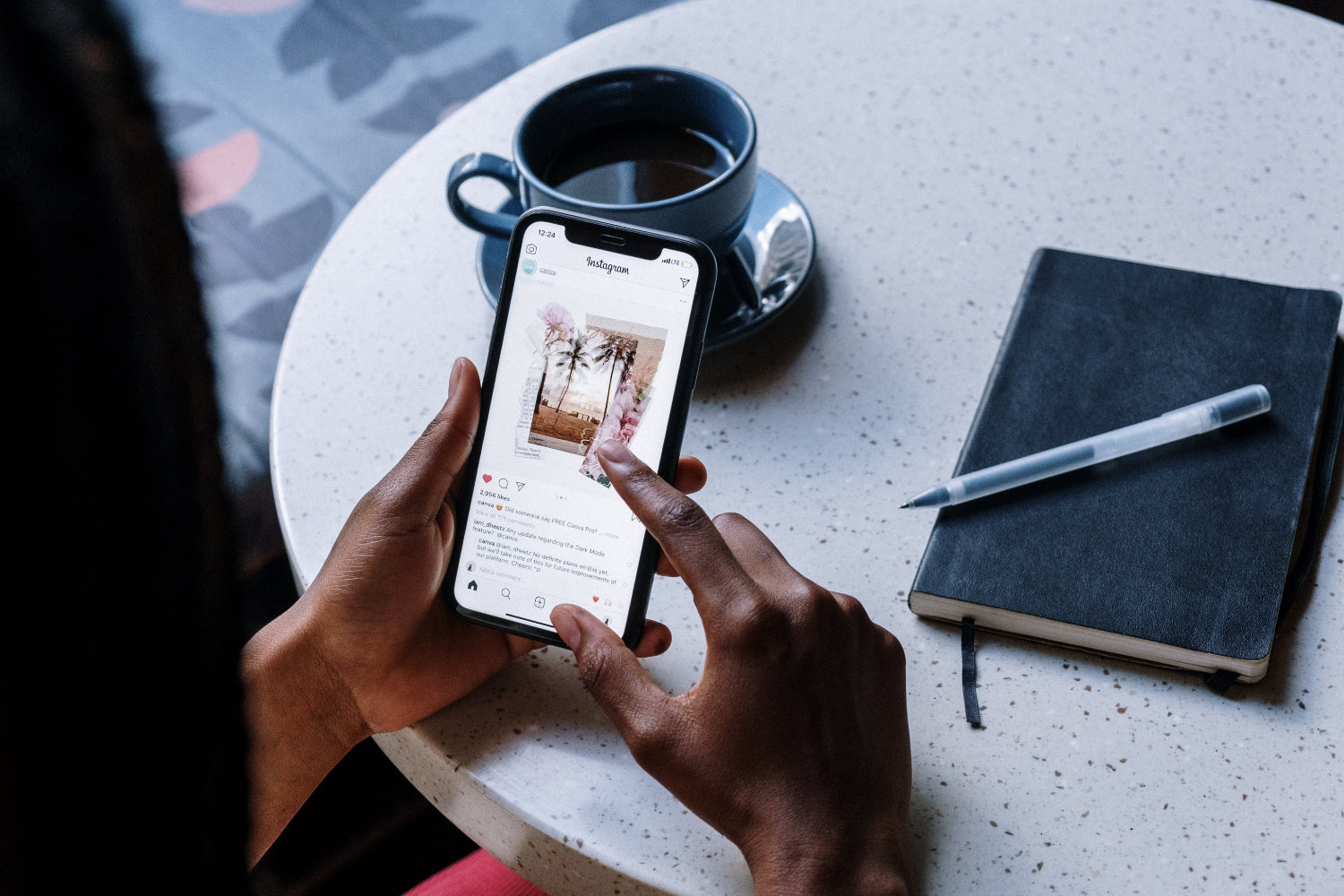 Person scrolling through Instagram on a smartphone at a table with a cup of coffee and a notebook.