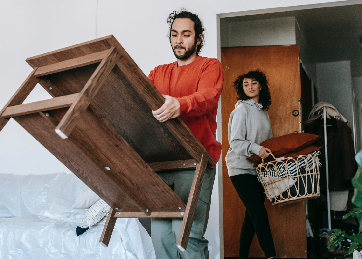 Man carrying a wooden table while a woman carries a basket of pillows into a room.