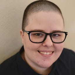 A person with a shaved head and black glasses smiles at the camera. They have a dark blue collared shirt on.