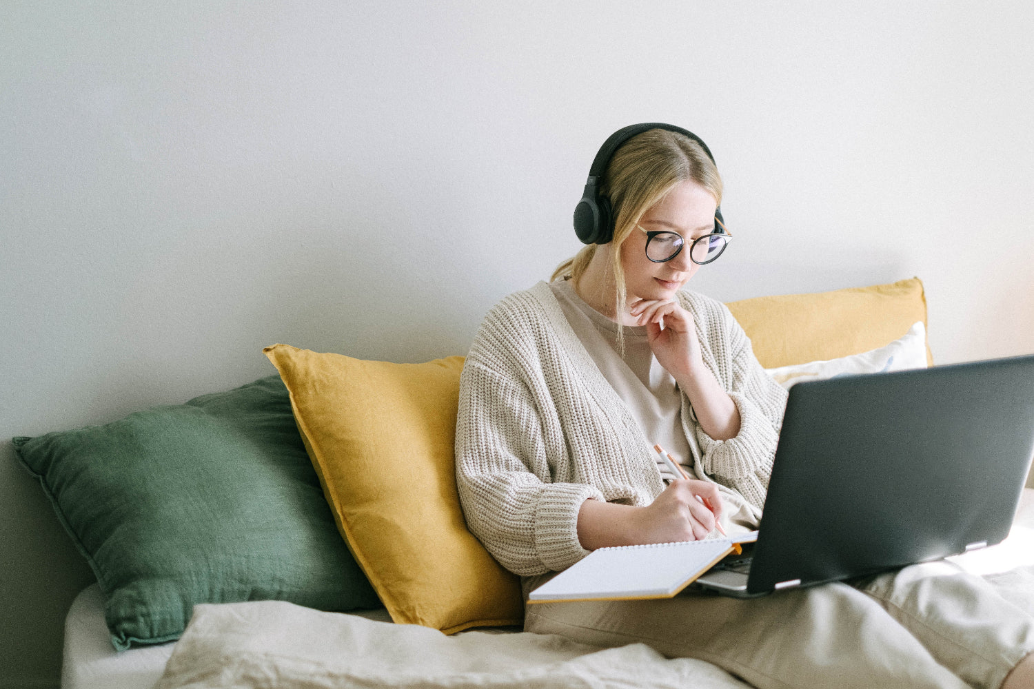 Woman with headphones writing in a notebook while using a laptop