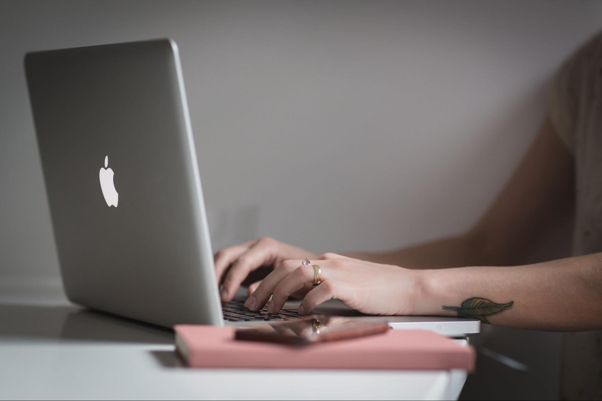 Close-up of hands typing on a MacBook with a notebook and phone nearby.